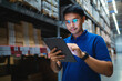 © KANGWANS - A man wearing glasses is looking at a tablet in a warehouse. He is focused on the screen and he is working on something