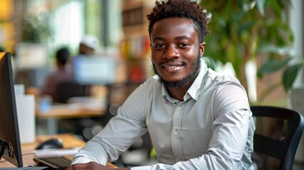 Poster - A young man with curly hair sits at a desk in a contemporary office space, smiling confidently while working on a computer. Natural light floods in from large windows.