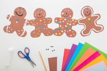 Wall Mural - Brown gingerbread man shapes created from paper. Scissors, glue stick, pencil and colorful application paper on light gray table background. Making decoration elements. Closeup. Top down view.