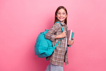 Wall Mural - Smiling young schoolgirl carrying a blue backpack and books against a pink background, wearing trendy outfit, exuding joy