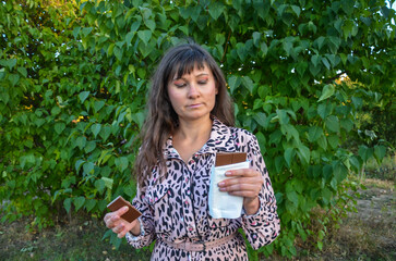 Woman stands in a lush garden, enjoying a chocolate bar. Her content expression highlights a love for sweets amidst serene natural surroundings, emphasizing relaxation and indulgence