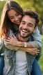 © natakot - A happy couple enjoys a sunny day in a lush green park, with the woman playfully hugging the man from behind. Their radiant smiles and carefree expressions capture the essence of love and joy