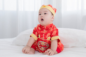 Wall Mural - Chinese New Year oriental culture celebration concept, adorable Asian boy in red costume smiling to camera, curiosity baby lying down in bed, Chinese infant in traditional clothes crawling development