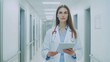 © vadosloginov - Professional female doctor confidently walking in a modern hospital corridor while reviewing a digital tablet during working hours
