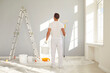 © Studio Romantic - Young man painter from repair service in white clothes standing back in empty room and holding a bucket with paint roller going painting a wall. Renovation and renovating house concept.