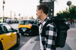 © BullRun - Rear view of caucasian male in trendy wear with backpack exploring city waiting for cab for transportation, 20s hipster guy standing on urban setting looking at cars and traffics in megalopolis