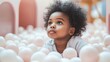 © LIDIIA - Dreamy Ball Pit Portrait. Young child with natural afro hair gazes upward while surrounded by soft pink and white balls in playful setting.