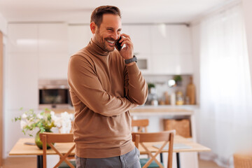 Wall Mural - Portrait of smiling handsome freelancer talking over mobile phone while standing in modern apartment