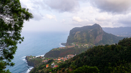 Wall Mural - A breathtaking coastal view of a hillside village surrounded by lush greenery and steep cliffs, overlooking the ocean under a cloudy sky. A serene blend of nature and settlement.