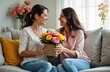 © Maryna - Two women sit on couch indoors. One woman gives bouquet of flowers to. Smiling, looking at. Scene suggests special occasion like Mothers Day. Casual attire suggests relaxed, happy atmosphere at home.