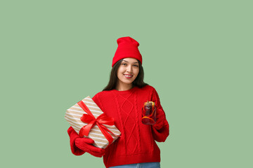 Wall Mural - Beautiful young happy Asian woman in winter clothes with glass jar of hot mulled wine and gift box on green background