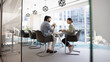 © fizkes - Two male and female coworkers talking at large conference table in contemporary office interior, meeting for teamwork, creative planning. Wide banner shot through glass doorway