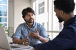 © fizkes - Confident Indian business project leader man talking to African partner workplace table, explaining task to manager, discussing teamwork, sitting at desk with laptop, speaking to colleagues