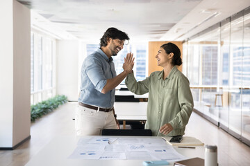 Happy couple of Indian business colleagues giving high five at workplace table with statistic paper reports, celebrating successful marketing analysis, enjoying teamwork, smiling, laughing