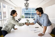 © fizkes - Positive satisfied young Indian business partners shaking hands over large meeting table, enjoying teamwork, friendship, celebrating successful startup, closing deal with handshake. Side view