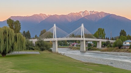 Wall Mural - Modern cable-stayed bridge over river with mountains in the background at sunset. Lush greenery and parkland surround the structure.