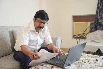 mature indian man working on laptop and checking document in a calm home setting. businessman and work from home concept.