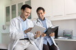 © fizkes - Two positive medical colleagues discussing electronic report, reading records on tablet, smiling, laughing. Happy Latin doctor consulting coworker, showing and pointing at gadget, asking for advice