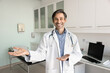 © fizkes - Happy positive successful Latin doctor man holding copy space on palms, pointing hands away, looking at camera with toothy smile, posing for promotion portrait in clinic examination office