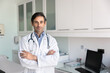 © fizkes - Serious Latin doctor man looking at camera for portrait, keeping arms folded, standing at workplace with desktop in hospital office, examination room, promoting successful medical career