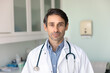© fizkes - Serious Latin middle aged doctor man in white uniform coat with stethoscope standing in clinic office, lab, examination room, looking at camera, posing for general practitioner, physician portrait