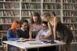© fizkes - Preparing for exams. Concentrated diverse polyethnic young people students pupils group gather around desk in library collaborate in studying books search answers on test questions do research discuss