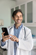 © fizkes - Happy middle aged Hispanic practitioner doctor man using mobile phone in his clinic office, typing, looking away, smiling, thinking on online communication with patients. Vertical shot