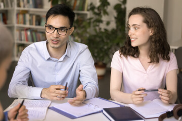 Wall Mural - Knowledge sharing. Smiling teen female international college university student take part in group conversation at library listen to young male groupmate in glasses speaking proposing idea to discuss