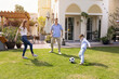© fizkes - Happy young parents and son kid kicking soccer ball on grass at house, playing football outside together, spending family leisure time outside, enjoying outdoor activities on backyard