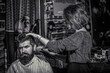 © Yevhen - Client during beard shaving in barbershop. Female barber at salon. Woman in the male profession. Barber cutting hair with clipper. Master provides service. Black and white