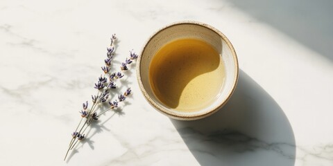 A close-up of a simple ceramic teacup holding herbal tea, paired with a sprig of lavender, with shadows creating a minimalist aesthetic on a marble surface