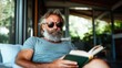 ©  Shomixer - A mature bearded man in sunglasses, seated comfortably indoors, deeply engrossed in a book, enjoying a moment of peace and reflection. Perfect for relaxation themes.