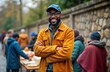 © Viktor - African man volunteer stands outdoors smiling at camera. Arms crossed. Homeless people receive food donations in urban area. Support, care shown during daytime. Community sharing, social aid. Charity