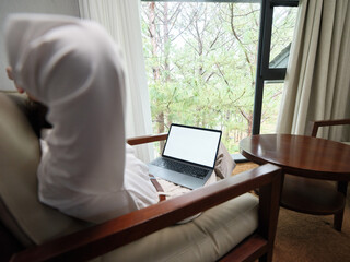 Canvas Print - Relaxed man working on laptop in cozy hotel room with natural light, green trees outside, emphasizing comfort and productivity while traveling