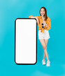 © sitthiphong - Cheerful and beautiful Asian woman stands beside a large smartphone mockup with a blank screen against a blue background.