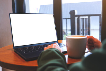 Wall Mural - Mockup image of a woman typing on laptop computer with blank white desktop screen while drinking coffee in cafe