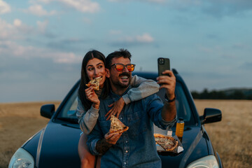 Wall Mural - Couple sharing pizza while leaning on a car and taking a selfies during a picturesque sunset