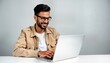 © Viktor - Young Indian man sits at table using laptop. He smiles happily while working. Modern workplace indoors. Casual attire. Virtual work. Technology lifestyle. Indoors. Happy man using computer.