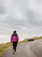 Wall Mural - Girl in colorful jacket walking on a small narrow road, Vast ocean view and cloudy sky on her left. Travel and tourism theme. Enjoy nature on your own.