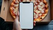 © Ashi - Person holding smartphone with blank screen above pizza in cardboard box, concept of online food ordering and delivery, artificial intelligence-generated visual