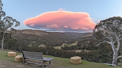 Wall Mural - Panoramic view of a valley with a pink cloud at sunset, featuring a wooden bench and hay bales.