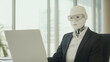© pattozher - The Rise of AI in Business: A humanoid robot in a business suit sits at a desk, working on a laptop, symbolizing the integration of artificial intelligence into the modern workplace.