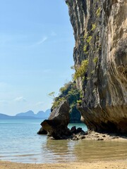 Poster - View of a rocky cliff by a beach with clear blue waters and distant islands under a bright sky.