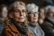 © Vilaysack - A thoughtful elderly woman with glasses gazes into the distance, surrounded by others, in a warm and inviting setting.