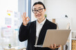 © sofiko14 - Confident Asian businesswoman in formal wear holding laptop, making okay gesture. Young female professional expressing approval and success in modern office environment.