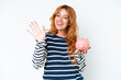 © luismolinero - Young caucasian woman holding a piggybank isolated on white background saluting with hand with happy expression