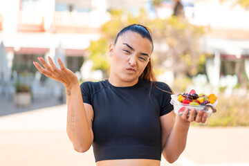 Wall Mural - Young pretty brunette woman holding a bowl of fruit at outdoors making doubts gesture while lifting the shoulders