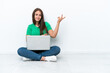 © luismolinero - Young Ukrainian woman with a laptop sitting on floor extending hands to the side for inviting to come
