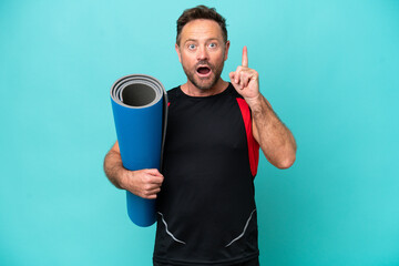 Wall Mural - Middle age sport man going to yoga classes while holding a mat isolated on blue background thinking an idea pointing the finger up