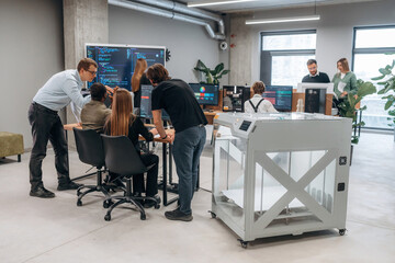 Big screen with code on the wall. People are in the office by the 3d printer